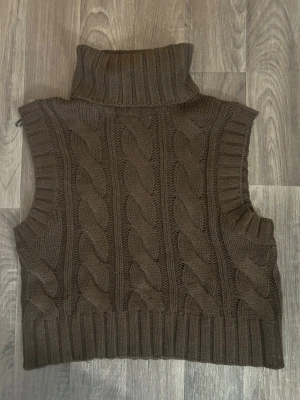 Brun stickad poloväst med flätmönster - Mysig brun stickad väst med hög polokrage och grovt flätmönster på framsidan. Västen har ribbade kanter vid ärmhål och nederkant, vilket ger en snygg och avslappnad look. Perfekt att bära över en skjorta eller tröja för extra värme.
