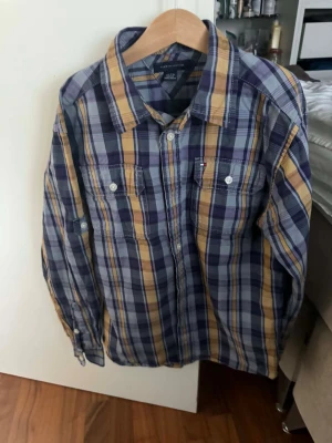 Rutig skjorta från Tommy Hilfiger - Snygg rutig skjorta från Tommy Hilfiger i lop blå, gul och vit. Skjortan har klassisk krage, två bröstfickor med knappar och långa ärmar. Tillverkad i mjuk bomull som känns skön mot huden. Perfekt för en avslappnad men ändå stilren look.