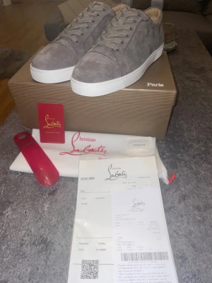 Gråa sneakers från Christian Louboutin - Snygga gråa sneakers från Christian Louboutin i mocka med klassisk röd sula och vita kanter. Skorna har lågt skaft, matchande grå skosnören och diskret branding på sulan. Perfekta för dig som gillar exklusiv och stilren design.