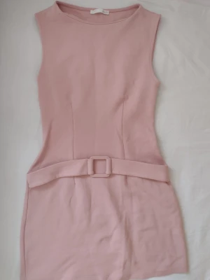 Ljusrosa ärmlös klänning med bälte - Säljer en stilren ljusrosa kortklänning med markerad midja och matchande bälte. Klänningen är ärmlös och har en enkel, elegant design som ger en feminin siluett. Perfekt för dig som gillar minimalistisk stil och mjuka färger.