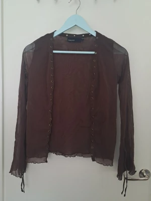 Brun meshkofta - Säljer en brun transparent meshkofta från KappAhl med långa ärmar och dekorativa öljetter längs öppningen. Koftan har tunna band vid ärmsluten och vågig kant nedtill. 🩷 str. 38/40