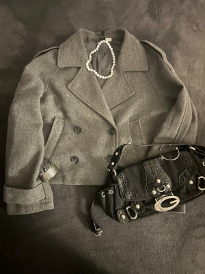 Grå dubbelknäppt ullrock - Kort grå rock i ullmix med bred krage och dubbelknäppning framtill. Rocken har axelklaffar, två sidofickor och dekorativa spännen vid ärmsluten. Klassisk och stilren modell som passar till många looks.