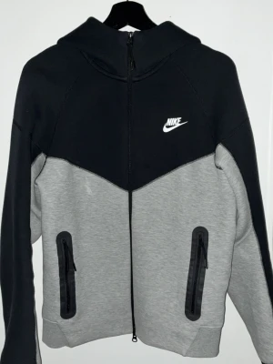 Nike tech hoodie nästan ny - Snygg hoodie från Nike i svart och grått med hel dragkedja framtill. Hoodien har två fickor med dragkedja och en vit Nike-logga på bröstet. Tillverkad i mjukt material och har en huva för extra komfort. Perfekt för en sportig och avslappnad stil kom privat för frågor.