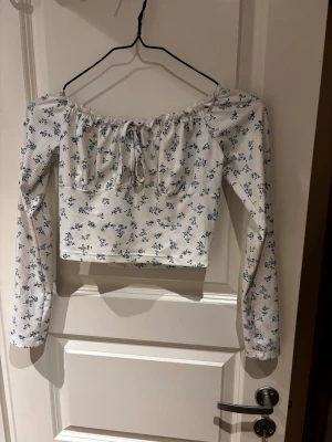 Blommig offshoulder topp  - Söt offshoulder topp med långa ärmar och blommigt mönster i blått på vit botten. Toppen har en croppad passform och knyt framtill vid urringningen. 