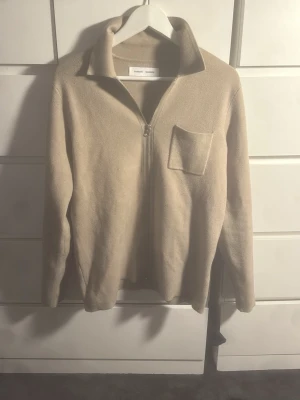 Beige ziptröja från Samsøe Samsøe - Stilren beige ziptröja från Samsøe Samsøe med krage och bröstficka. Tröjan har lång ärm och är tillverkad i ett mjukt material som känns skönt mot huden. Perfekt för lager på lager och enkel att matcha med olika outfits.