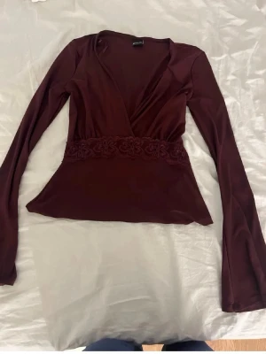 Vinröd omlottopp med spetsdetalj - Snygg vinröd omlottopp med långa ärmar och v-ringning. Toppen har en bred spetsdetalj i midjan med blommönster som ger en extra touch. Tillverkad i ett mjukt, stretchigt material som sitter bekvämt på kroppen.