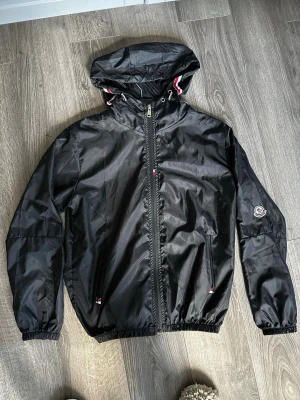 Svart vindjacka från Moncler - Snygg svart vindjacka från Moncler med huva och dragkedja framtill. Jackan har elastiska muddar, två fickor och Moncler-logga på ärmen. Detaljer i rött och vitt vid huvan ger en sportig touch. Tillverkad i lätt, glansigt material. Pris kan diskuteras 