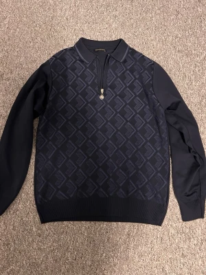 Mörkblå tröja från Stefano Ricci - Snygg mörkblå tröja från Stefano Ricci med diskret geometriskt mönster på framsidan. Tröjan har halv dragkedja med metall-detalj och ribbade muddar. Perfekt för dig som gillar exklusiv och stilren design. Är villig att komma överens om ett pris