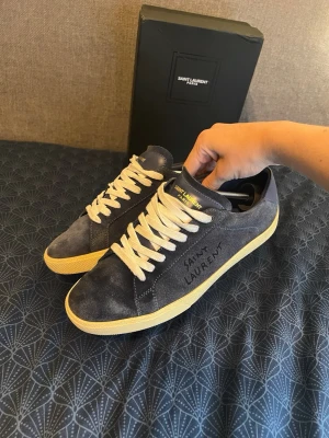 Saint Laurent blå mocka sneakers - Mörkblå sneakers från Saint Laurent i mjuk mocka med vita snören och gulaktig sula. Klassisk låg modell med Saint Laurent-logga broderad på sidan och diskret branding på plösen. Perfekta för en clean och stilren look.