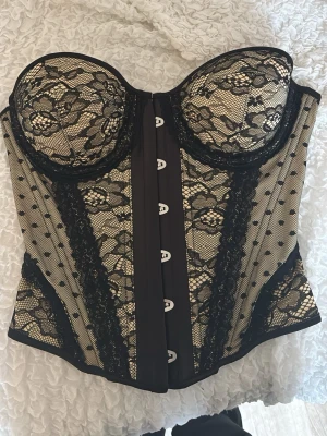 Svart spetskorsett med mönster - Säljer en snygg korsett i svart spets med blommigt mönster och beige undertyg. Korsetten har vadderade kupor, hakknäppning fram och dekorativa detaljer i spets och mesh. Perfekt för dig som vill ha en elegant och feminin look. Köptes av en tjej här på plick och lappen verkar vara bortklipp därav vet jag inte vart den är köpt