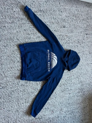 Blå H&M hoodie med Global Squad text - Mörkblå hoodie från H&M med Global Squad med vit logga och text på bröstet. Klassisk känguruficka framtill och huva. Tillverkad i mjukt bomullsmaterial, perfekt för chill dagar. Långärmad och enkel design utan dragkedja.