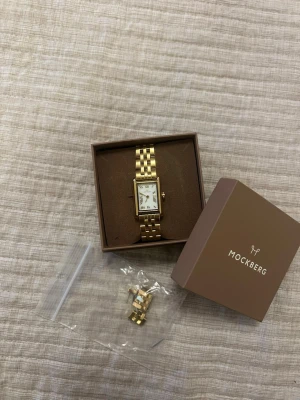 Mockberg guldig klocka med länkarmband - Snygg guldig klocka från Mockberg med rektangulär urtavla och klassiskt länkarmband. Urtavlan är vit med romerska siffror och gulddetaljer. Kommer i originalask och extra länkar medföljer. Perfekt accessoar för en stilren look. Den är i bra skick men har lite färgslitage på låset, men syns inte när man spänner fast den.