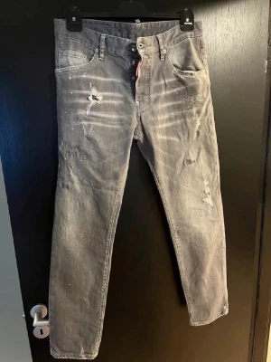 Gråa Dsquared2 Jeans - Säljer ett par grå jeans från DSQ2 med slitna detaljer, små hål och färgstänk för en trendig, avslappnad look. Klassisk femficksmodell med rakt ben och knappgylf. Perfekta för dig som gillar streetwear och en edgy stil. Storlek 50