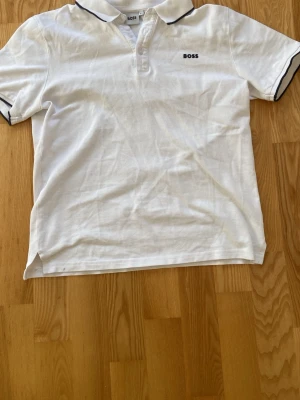 Vit pikétröja från BOSS - Klassisk vit pikétröja från BOSS med svart logga på bröstet och svarta detaljer vid krage och ärmslut. Tillverkad i mjuk bomull för skön känsla. (Nypris 1000kr)