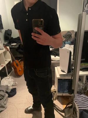 Svart pikétröja från Ralph Lauren helt ny - Klassisk svart pikétröja från Ralph Lauren med röd broderad logga på bröstet. Tröjan har korta ärmar, krage och tre vita knappar framtill. Perfekt till jeans eller shorts för en clean och stilren look. Helt ny! Oanvänd!