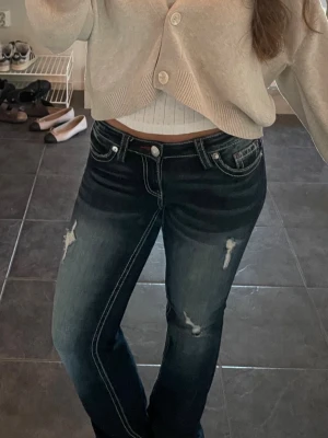 Mörkblå low bootcut jeans med slitningar - Snygga mörkblå bootcut jeans med low waist och slitna detaljer på låren och knäna. Jeansen har vita kontrastsömmar, klassisk femficksdesign och låg midja. Perfekta för en avslappnad och trendig look.