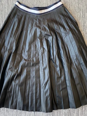 Svart plisserad långkjol med resår i skinn look - Snygg svart långkjol med plisserade veck och bred resår i midjan med vit och marinblå rand. Kjolen har en klassisk och stilren look som passar till många olika outfits. Materialet känns mjukt och följsamt.Svart plisserad kjol i skinnlock i strl 6 (36) fr Tommy Hilfiger   Endast använd vid ett par tillfällen och är i nyskick  Mått Midjan: ca 36 cm Total längd: ca 68 cm  47% polyester, 43% polyvonyl och 10% polyuretan   Kommer från ett djur och rökfritt hem
