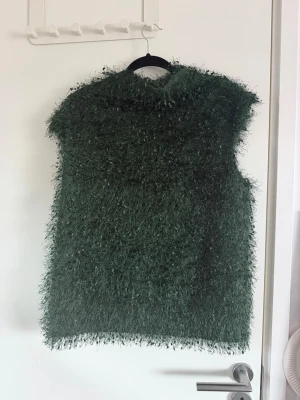 Mörkgrön fluffig tröja - Unik mörkgrön tröja med fluffig och långhårig fuskpäls. Tröjan har rak passform och är ärmlös, perfekt för dig som vill sticka ut med en cool och trendig look. Materialet är syntetiskt och ger en mjuk känsla. Inga synliga fickor eller knäppning.