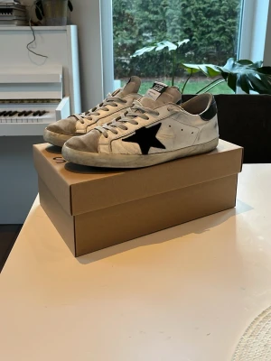 Golden Goose superstar vita sneakers med svart stjärna - Golden Goose Superstar sneakers i vitt skinn med grå mockadetaljer och svart stjärna på sidan. Skorna har svarta hälpartier, snörning och platt sula. Klassisk streetstil med GGDB-logga på plösen och hälen. Perfekta för dig som gillar en avslappnad och trendig look. OBS originalboxen tillkommer ej! Bara att skriva vid funderingar!