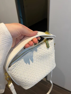 Vit axelremsväska med ormmönster - Säljer en snygg vit handväska med axelrem och guldfärgade detaljer. Väskan har ett stilrent ormmönstrat yttre i konstläder och dragkedja upptill. Perfekt för dig som gillar trendiga accessoarer och vill ha något som sticker ut.