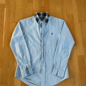 Ralph Lauren skjorta - Klassisk ljusblå skjorta från Ralph Lauren med button-down krage och rutigt innerfoder i kragen. Skjortan har lång ärm, knappar framtill och den ikoniska broderade loggan på bröstet. Perfekt för en clean och stilren look.