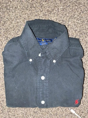 Svart skjorta från Ralph Lauren - Svart skjorta från Ralph Lauren, slim fit. Rätt bra använd, 6/10 skick. Inga större defekter men lite naturlig "missfärgning" här och där av tvätten. Inget som sticker ut när man har på sig den.