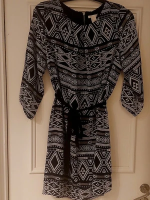 Svartvit mönstrad kortklänning - Svartvit kortklänning med grafiskt mönster och trekvartsärm. Klänningen har ett knytband i midjan och rund halsringning. Lätt och luftig modell som ger en avslappnad men snygg look.