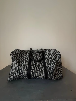 Svartvit Dior weekendbag canvas (EJ ÄKTA)  - Säljer en svartvit Dior weekendbag i canvas med det klassiska Dior-monogrammönstret över hela väskan. Väskan har svarta handtag i läder och silverfärgade metalldetaljer. Perfekt för resor när du vill ha både stil och funktion. Den är inte äkta, men helt oanvänd och nyskick! 