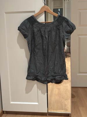 Mörkgrå blus med smockdetaljer - Snygg mörkgrå blus med korta ärmar och smockade partier vid axlarna. Blusen har rund halsringning med volangkant och lätt rynkade detaljer både upptill och nertill. Tillverkad i ett tunt, mjukt tyg som ger en avslappnad look.