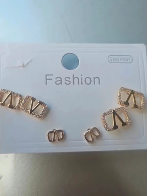 Guldiga örhängen med V-logga - Säljer ett set med guldiga örhängen i olika storlekar, alla med en stilren V-logga. Några av örhängena har gnistrande stenar runt kanten för extra bling. Materialet är metall och vissa delar är dekorerade med små kristaller.