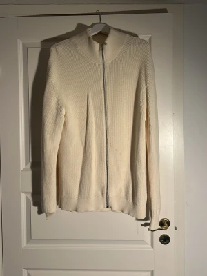 Vit ribbstickad ziptröja - Klassisk vit ribbstickad tröja med hel dragkedja framtill och hög krage. Tröjan har lång ärm och är tillverkad i ett mjukt, tjockare material som ger en clean och stilren look. Perfekt för lager på lager och enkel att matcha.