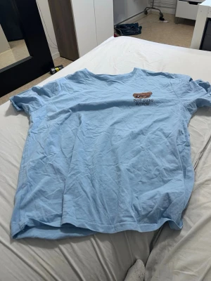 Ljusblå t-shirt med björntryck - Ljusblå t-shirt med ett tryck av en björn med solglasögon och texten 'Busy doing nothing...' på bröstet. T-shirten har en avslappnad passform och är kortärmad, perfekt för en chill och casual stil. Tillverkad i mjuk bomull.