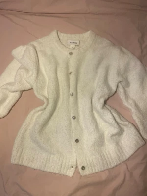 Vit fluffig kofta från Monki - Mysig vit kofta från Monki med fluffig textur och rund halsringning. Koftan har långa ärmar, knappar framtill i pärlemor och ribbade muddar vid ärmslut och nederkant. Perfekt för lager på lager och en clean look.