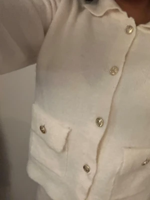 Vit kofta med guldknappar - Stilren vit kofta med stora guldknappar och två fickor framtill. Koftan har en klassisk krage och långa ärmar med breda muddar. Perfekt för en chic och trendig look.