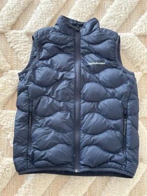 Mörkblå dunväst Peak Performance - Snygg mörkblå dunväst från Peak Performance med hög krage och två dragkedjeförsedda fickor. Använd ett fåtal gånger. 
