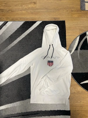 Vit hoodie från GANT med logga - Vit hoodie från GANT med broderad logga och sköld på bröstet. Klassisk huva med svarta snören och känguruficka framtill. Perfekt för en chill och stilren look.