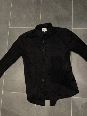 Svart skjorta - Klassisk svart skjorta från H&M No 6 med lång ärm och knappar framtill. Skjortan har en enkel, stilren design och är tillverkad i bomull vilket gör den bekväm och lätt att bära. Perfekt för dig som gillar en clean och tidlös look.