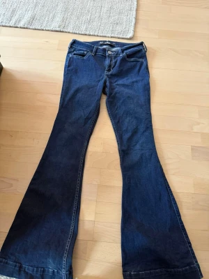 Mörkblå bootcut jeans med broderi - Säljer ett par mörkblå bootcut jeans med snygga vita broderade detaljer på bakfickorna. Jeansen har klassisk femficksmodell och är tillverkade i ett mjukt denimtyg. Perfekta för dig som gillar retrovibbar och vill ha ett par jeans med lite extra attityd.