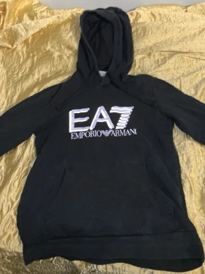 Svart EA7 Emporio Armani hoodie - Snygg svart hoodie från EA7 Emporio Armani med stor vit logga på bröstet. Klassisk huva med dragsko och magficka framtill. Perfekt för en avslappnad och trendig stil. Lite dåligt skick men värt för priset!