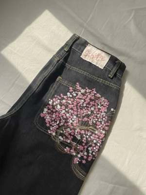 Y2K Black Denim Jeans with Pink Cherry Blossom Embroidery - streetwear style - Very clean black denim jeans with detailed pink cherry blossom embroidery on the pocket. A rare and eye-catching design that gives strong Y2K and streetwear vibes while still being easy to wear daily.  High-quality denim, sturdy stitching, and comfortable fit. The embroidery is in excellent condition with no flaws.  Perfect for streetwear lovers, Y2K outfits, alternative fashion, or anyone looking for unique embroidered jeans that stand out. Easy to style with hoodies or graphic tees