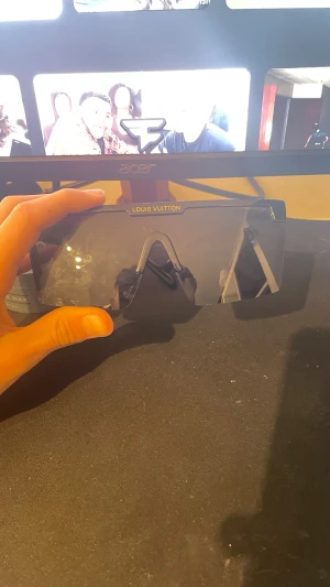 Louis Vuitton svarta shield-solglasögon - Säljer ett par svarta shield-solglasögon från Louis Vuitton med bred, futuristisk design och logotyp på linsen. Bågarna är i blank svart plast och har Tom Walker-signatur på sidan. Perfekt för dig som vill sticka ut med en lyxig accessoar.