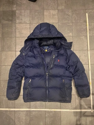  Mörkblå dunjacka Polo Ralph Lauren  - Snygg mörkblå dunjacka från Polo Ralph Lauren med röd broderad logga på bröstet. Jackan har huva, flera dragkedjeförsedda fickor och RL-detaljer på zipparna. Puffad design och långärmad, perfekt för kalla dagar.