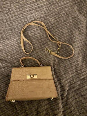 Brun axel/hand väska - Snygg brun axel/hand väska. Väskan har guldfärgade metalldetaljer, ett topphandtag och en avtagbar axelrem. Klassisk fyrkantig form och vridlås framtill. Perfekt för dig som gillar trendiga accessoarer.