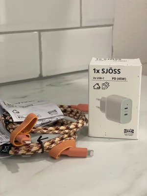 IKEA SJÖSS USB-C laddare & kabel - Säljer en IKEA SJÖSS väggladdare med 2x USB-C-portar (45W PD) och en snygg flätad laddningskabel med orange och bruna detaljer. Perfekt för att ladda mobilen snabbt och smidigt. Kabeln har USB-C till Lightning-kontakt och är klädd i slitstarkt tyg.