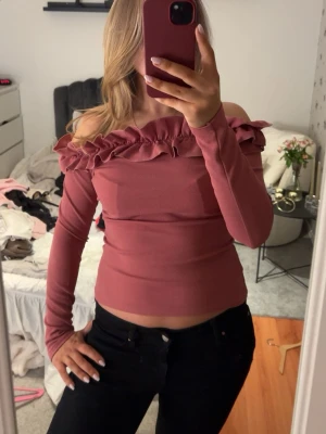 Rosa offshoulder topp med volang - Trendig rosa offshoulder topp med långa ärmar och volangdetalj längs halsringningen. Sitter tight och har en croppad passform som ger en snygg siluett. Perfekt för dig som vill sticka ut med en feminin och modern stil.