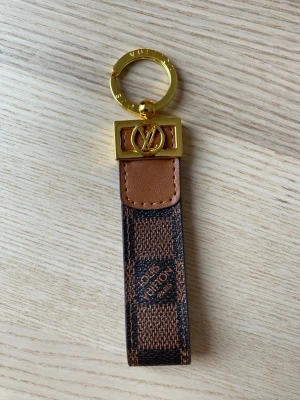  Vuitton nyckelring brun/guld - Snygg nyckelring från Vuitton i klassiskt brunt Damier-mönster med detaljer i guld och läder. Har en guldfärgad ring och LV-logga i metall. Perfekt accessoar för att piffa till väskan eller nyckelknippan.