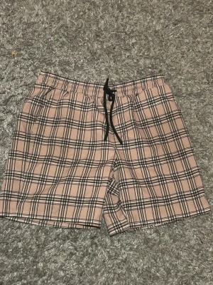 Burberry shorts med snörning  - Snygga rutiga shorts i beige och svart med elastisk midja och svart snörning framtill. Perfekta för sommaren den fungerar väll bra till simningen och har en avslappnad passform. Tillverkade i mjukt material som känns skönt mot huden.