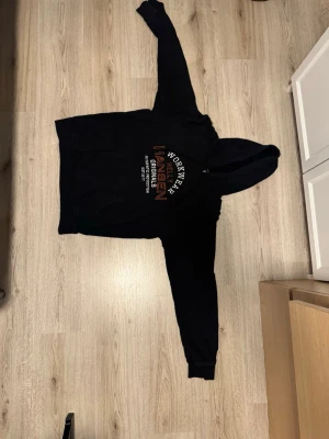 Svart hoodie från Worker Wear - Svart hoodie från Worker Wear med tryck på bröstet där det står 'Worker Wear Originals'. Klassisk modell med huva och känguruficka framtill. Mjuk insida och ribbade muddar vid ärmar och nederkant. Perfekt för en avslappnad stil.