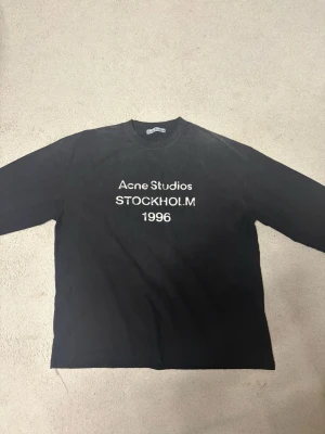 Svart långärmad topp Acne Studios - Svart långärmad topp från Acne Studios med vit text 'Acne Studios STOCKHOLM 1996' på bröstet. Klassisk rund halsringning och rak passform. Perfekt för en clean och stilren look.