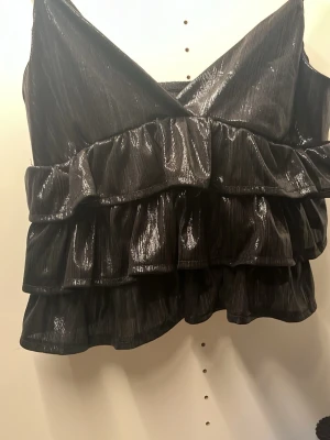 Glittrig svart volangtop - Snygg svart topp med glansigt tyg och flera volanger. Djup v-ringning och breda axelband ger en trendig look. Perfekt för dig som vill sticka ut med en cool och modern stil. Materialet är skimrande och ger ett exklusivt intryck. Det finns ingen lapp inuti så jag vet inte vart den är köpt då jag fick den av en kompis.
